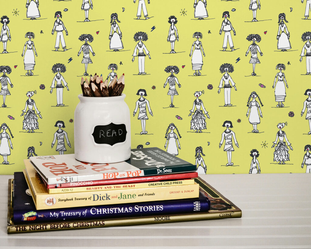 A bright yellow wallpaper featuring a repeating pattern of stylized, hand-drawn female figures wearing different outfits and hairstyles. The figures are in black and white with small colorful details. Accent wall idea for eccentric interior design!